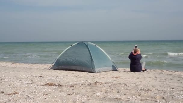 若い女性の観光客がテントの近くの海岸に座っている スマートフォンで写真を撮る レジャー — ストック動画