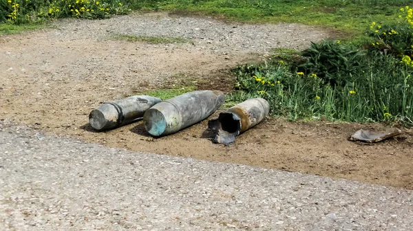Tři dělostřelecké granáty leží na kraji asfaltové cesty, pampelišky kvetou a tráva zelená — Stock fotografie