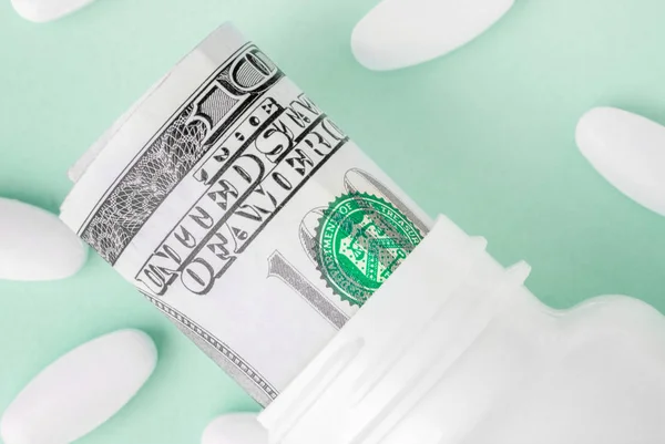 Roll of money Bills in white pill bottle with white oval pills around on Aqua Menthe background. medicine flat lay — Stock Photo, Image