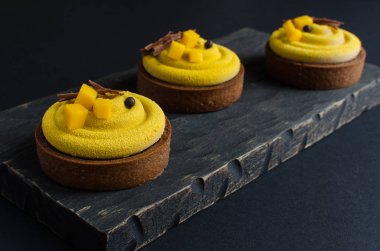 Çağdaş çikolatalar ve siyah arka planda taze mango ile süslenmiş sarı kadife spreyli mango tarları.