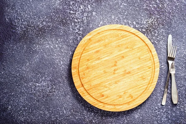 Empty round cutting board on a table with fork and knife, copy space at the side for your text. — Stock Photo, Image
