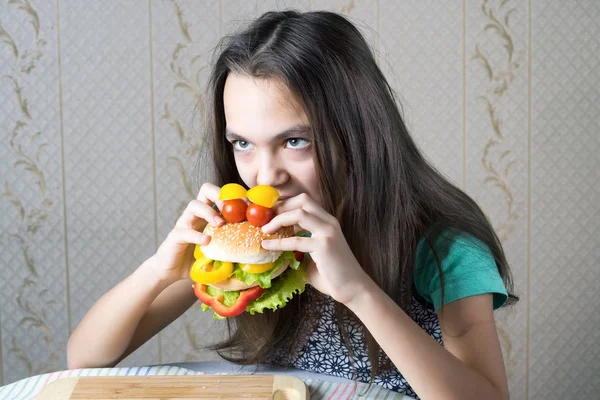 11 jaar oud meisje bijt haar tanden in een hamburger, gemaakt in de vorm van gezichten met ogen van cherry tomaten. — Stockfoto