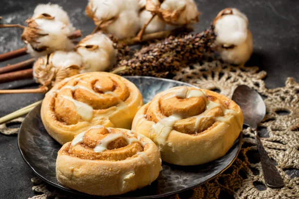 Three icing cinnamon rolls on a metal plate with spoon and crocheted doily on a dark cement background with cotton boxes behind. — Stock Photo, Image