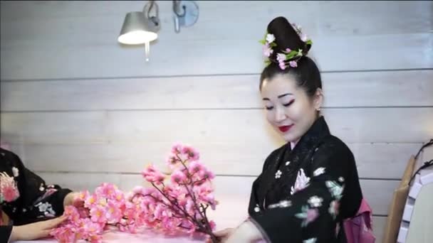 Hermosa geisha en kimono tradicional japonés sentado en una mesa vacía en el restaurante y recoge en las ramas de flores de cerezo . — Vídeos de Stock