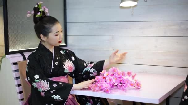 Hermosa geisha en kimono tradicional japonés sentado en una mesa vacía en el restaurante y recoge en las ramas de flores de cerezo . — Vídeos de Stock