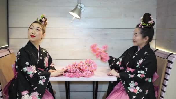 Dos hermosas geishas en kimono japonés tradicional sentadas en una mesa vacía en el restaurante y recoger en las ramas de flores de cerezo . — Vídeo de stock