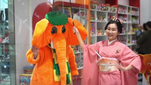 Moscú, Rusia - 5 de marzo de 2017: geisha en kimono tradicional japonés se para y abraza con un hombre en un traje de elefante naranja durante las actuaciones en el centro comercial "Oceanía" . — Vídeos de Stock