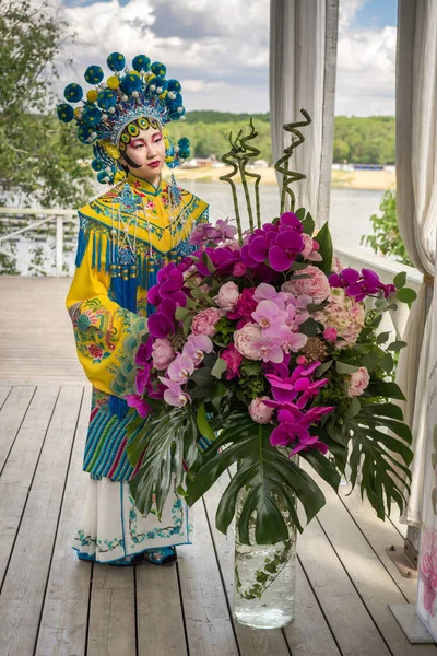 Όμορφη κοπέλα κινέζικα σε παραδοσιακά κινέζικα φόρεμα και pompon καπέλο στέκεται κοντά στο μεγάλο παιωνία και ανθοδέσμη ορχιδέα — Φωτογραφία Αρχείου