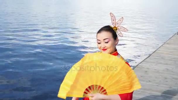 Belle geisha dans une robe traditionnelle chinoise rouge assis au bord de la rivière avec ventilateur jaune — Video