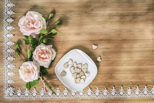 Disposición plana de rosas rosadas con platillo blanco y corazones de madera en una mesa de madera con marco de encaje y espacio de copia para su texto —  Fotos de Stock