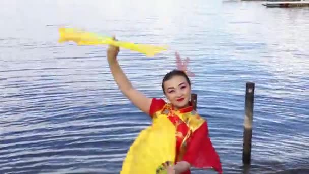 Hermosa mujer asiática en tradicional chino rojo kimono bailando con dos grandes fans amarillos en la orilla del río — Vídeo de stock