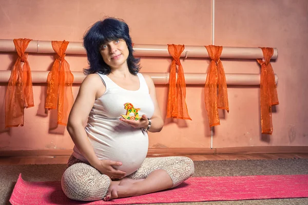 Gebelik Yoga ve Fitness kavramı. Sağlıklı annelik yaşam kavramı. küçük tahta at oyuncak ile yoga egzersiz yaparak lotus asana oturan 40 haftalık hamile orta yaşlı beyaz kadın.
