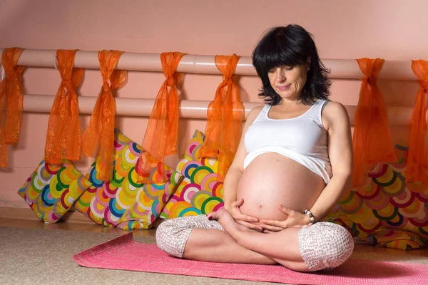 Pregnancy Yoga and Fitness concept. Healthy maternity lifestyle concept. 40 week pregnant middle aged caucasian woman sitting in lotus asana doing yoga exercises and hugging her stomach with softness.
