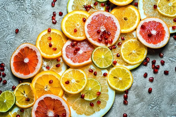 diagonal composition of slised citruses, lemon, lime, orange and pomegranate seeds on a rustic beige cement background, top view.