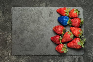 Kavram bireysellik, seçkin, daha iyi bir seçim. Siyah taş tepsi arka plan üzerinde kırmızı çilek arasında bir mavi çilek. Kopya alanı ile Üstten Görünüm.