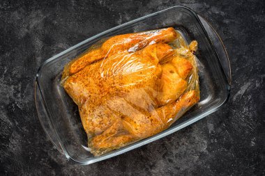 bir siyah çimento arka plan üzerinde yemek pişirmek için hazır bir cam tepsi fırın bir çanta içinde baharatlar ile ham bütün tavuk.