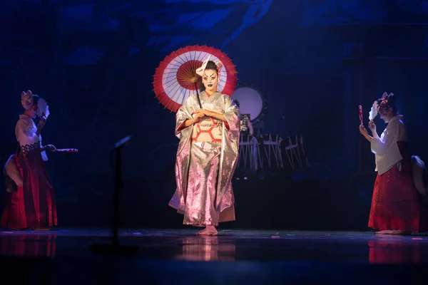 Traditional Japanese performance. Actress in traditional kimono and fox mark dancing with umbrella on a dark stage.