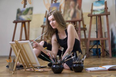 Sanat stüdyosundaki sanatçı, çelik tezgahın üzerine resim çiziyor, yerde oturuyor..