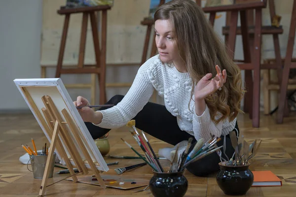 Woman artist sits on the floor in an art studio and paints an oil painting on canvas.