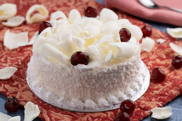 white forest cake with white chocolate shavings and cherries as topping placed on a red table cloth