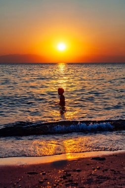 Çocuk arka planda inanılmaz bir günbatımıyla yüzerken eğleniyor. Sudaki çocuk bir siluet gibi