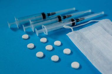 Yüzünde tıbbi maske, şırıngalar ve beyaz haplar var, mavi arka planda.