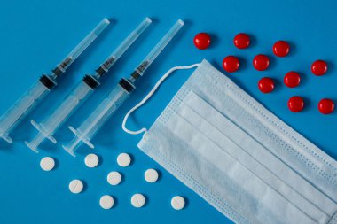 Yüzünde tıbbi maske, şırıngalar ve yukarıdan mavi arka planda beyaz ve kırmızı haplar var.