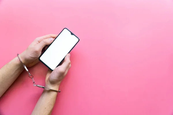 Elleri kelepçeli erkekler pembe arka planda boş ekranlı modern bir akıllı telefonu tutarlar. İnternet ve aygıt bağımlılığı kavramı.