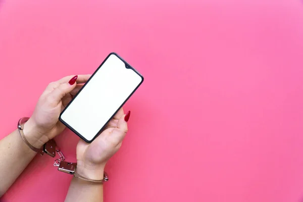 Elleri kelepçeli bir kadın pembe arka planda bir akıllı telefon tutuyor. İnternet ve aygıt bağımlılığı kavramı.