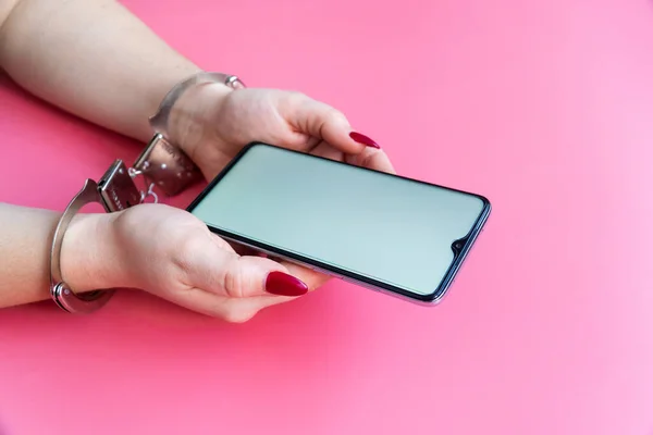 Kadın elleri kelepçeli, pembe arka planda modern bir akıllı telefon tutuyor. İnternet ve aygıt bağımlılığı kavramı.