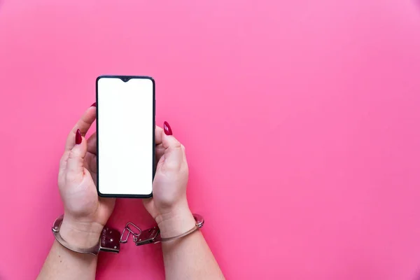 Kadın elleri kelepçeli, pembe arka planda boş ekranlı modern bir akıllı telefon tutuyor. İnternet ve aygıt bağımlılığı kavramı.