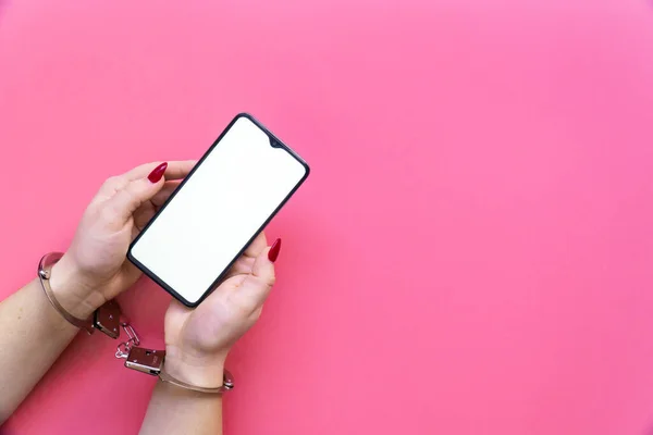 戴手铐的女人手持粉色背景的智能手机。互联网和电子产品依赖的概念. 图库照片