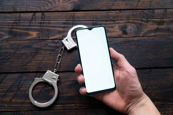 Mans mão segurar um smartphone moderno com uma tela em branco, algemas mentira contra um fundo de madeira — Fotografia de Stock