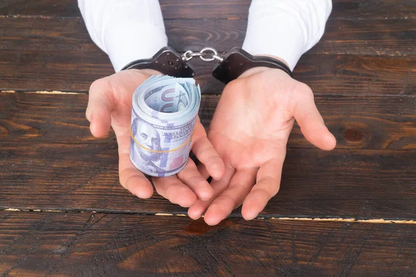 Elleri kelepçeli erkek eller, karanlık bir ahşap zemin üzerinde çarpık para tutar, rüşvet ve yolsuzluk kavramı