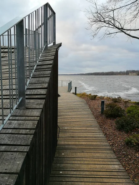 Wooden pier that leads to a viewing platform. Wooden site in front of the river. Wooden walk board with a fence at cloudy day. Pier with balcony.