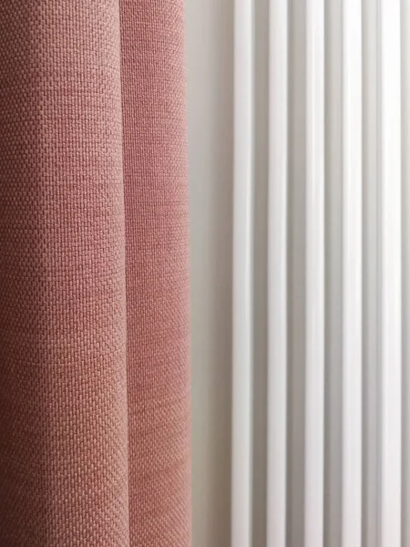 Folds of pink linen curtains in front of the white heating system. Abstract vertical lines in interior. Contrast between linen fabric and bright white metal in interior design.