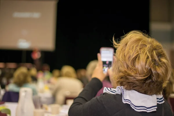 Woman using technology to help take in information while watching a keynote speaker at a conference with a slide deck presentation projected