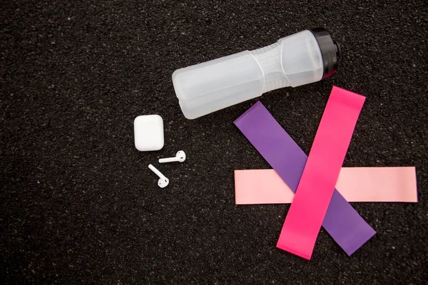 Three types of fitness gum rubber bands for sports, wireless headphones and a bottle of water on black background