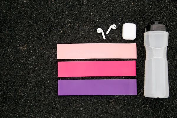 Three types of fitness gum rubber bands for sports, wireless headphones and a bottle of water on black background