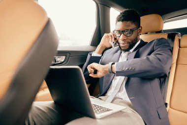 Araba sürerken yanında dizüstü bilgisayar olan bir adam telefonda konuşuyor..