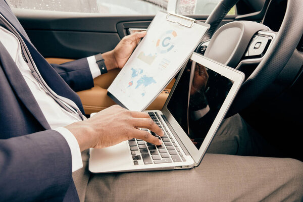 Man works in his car with laptop and notepad in front of him.