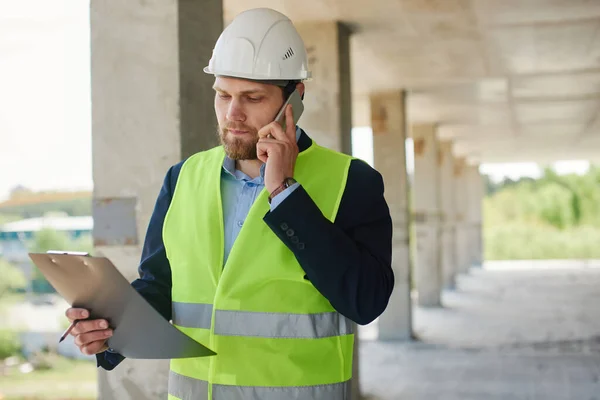 An engineer in a helmet talks on the phone and holds a notepad in a close-up.
