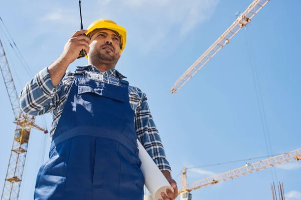 An engineer uses radio set on his work place with a folded draft.