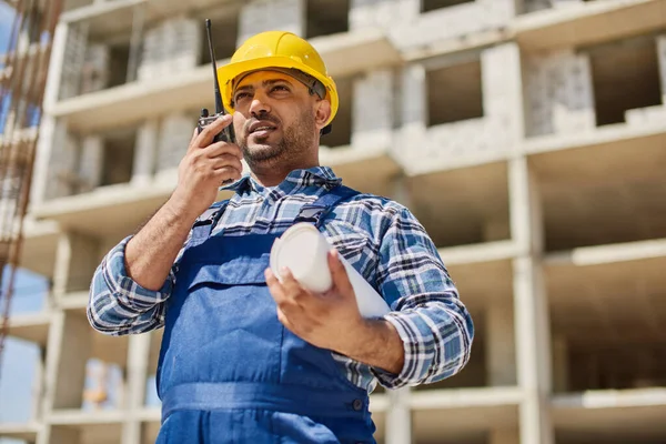 An engineer uses radio set on his work place with a folded draft.