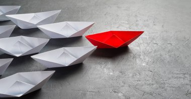 İş liderliği kavramı, Paper Boat, anahtar fikir lideri, nüfuz kavramı. Lider olarak bir kırmızı kağıt tekne, gri beton arka planda beyaz gemilere doğru gidiyor.