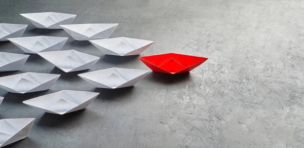 İş liderliği kavramı, Paper Boat, anahtar fikir lideri, nüfuz kavramı. Lider olarak bir kırmızı kağıt tekne, gri beton arka planda beyaz gemilere doğru gidiyor.