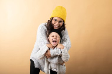 Genç ve enerjik bir anne, turuncu bir arka planda oğluna sarılıp onu öper.