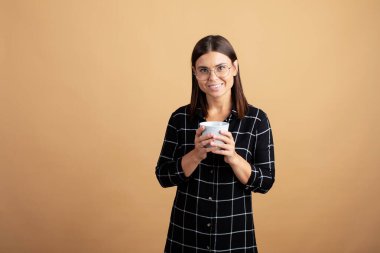 Ekose elbiseli genç bir kadın elinde mavi bir kupayla turuncu arka planda duruyor.