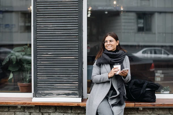Портрет молодой женщины в очках. Молодая деловая женщина стоит у окна кафе и пьет кофе — стоковое фото