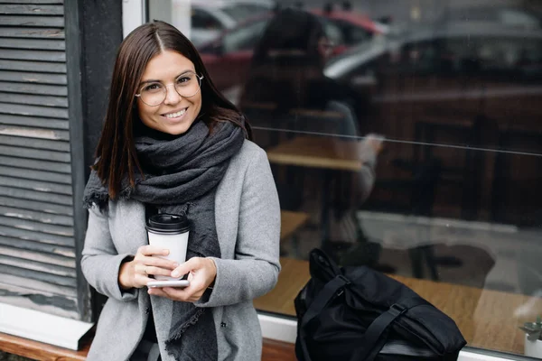 Портрет молодой женщины в очках. Молодая деловая женщина стоит у окна кафе и пьет кофе — стоковое фото